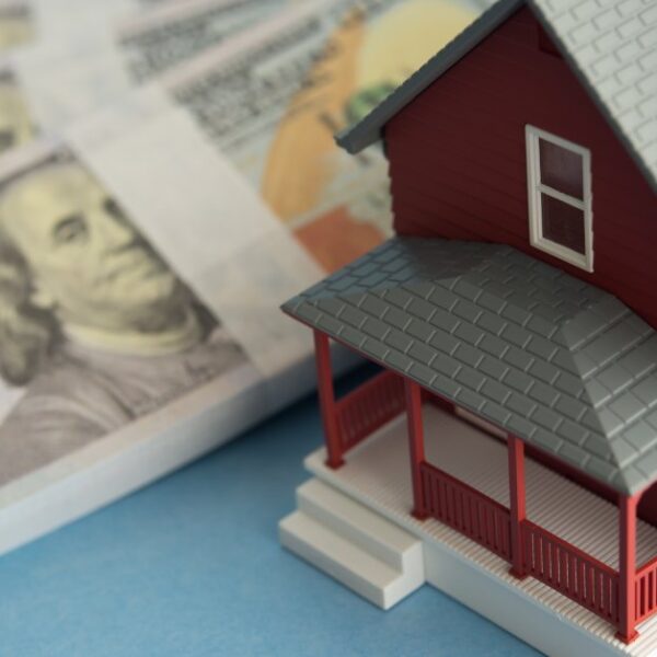 A close-up of a red model home with a gray roof sitting next to stacks of one-hundred-dollar bills on a table.