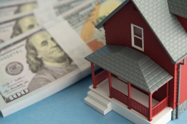 A close-up of a red model home with a gray roof sitting next to stacks of one-hundred-dollar bills on a table.