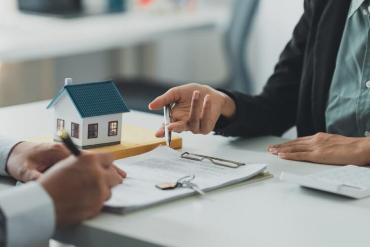 Two people wearing business attire sit across from each other at a desk with clipboards, documents, and a model home between them.
