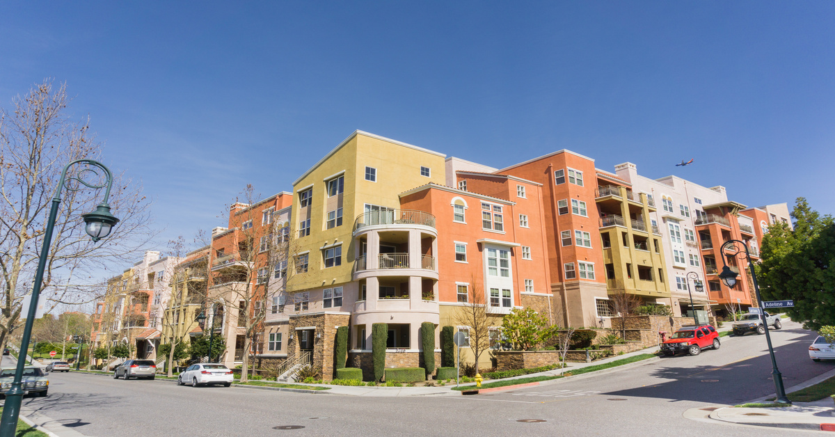 A corner view of a multifamily housing building with orange, yellow, and white siding and cars parked on the sides of the street.