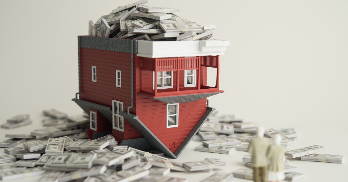 A red model home flipped over to hold bundles of 100-dollar bills, which are spilling out of the model's base.