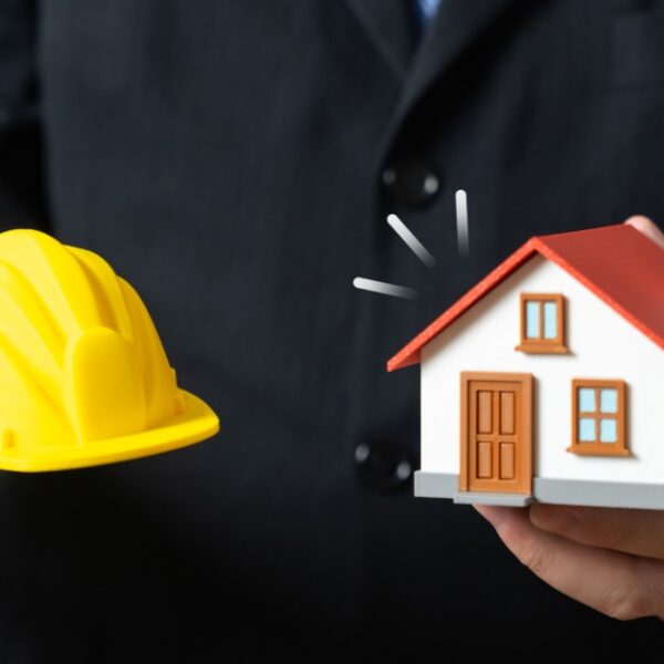 A close-up of a man in a black suit holding a model house in one hand and a mini hard hat in the other.