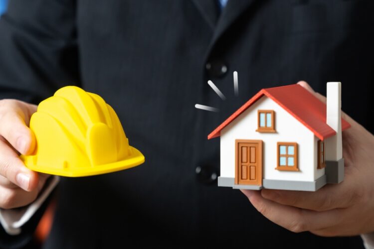 A close-up of a man in a black suit holding a model house in one hand and a mini hard hat in the other.