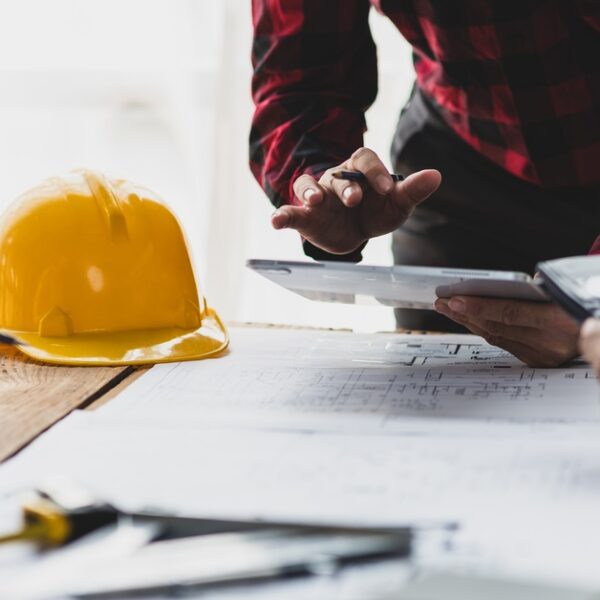 A man in a red, plaid shirt leans over blueprints and a hard hat on a table with a tablet in hand, two other people sitting across.