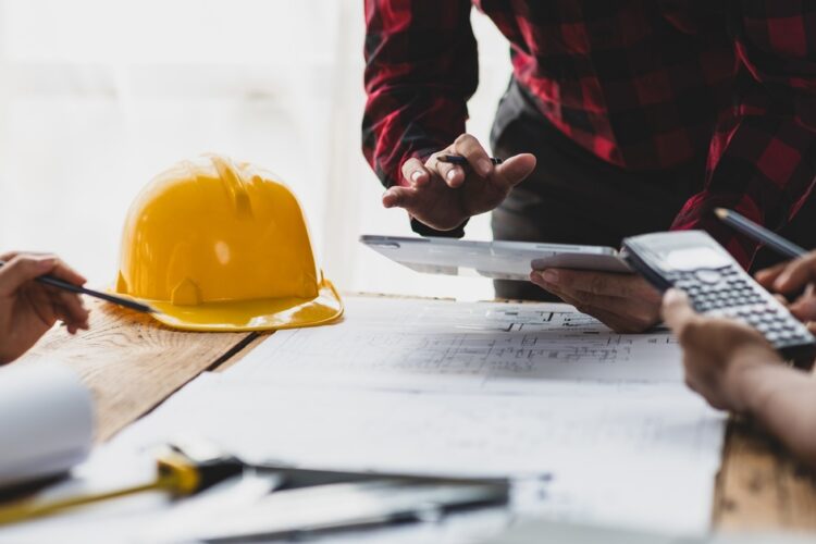 A man in a red, plaid shirt leans over blueprints and a hard hat on a table with a tablet in hand, two other people sitting across.