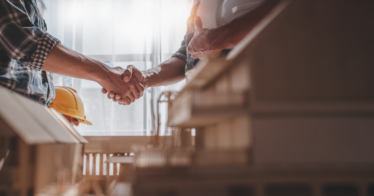 A handshake between two men, one holding a hard hat in the other hand. Two model homes sit in the blurry foreground.