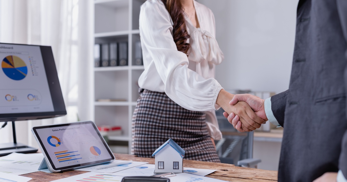 A businesswoman wearing nice clothes shakes hands with a man wearing a business suit over a desk wirh scattered documents.