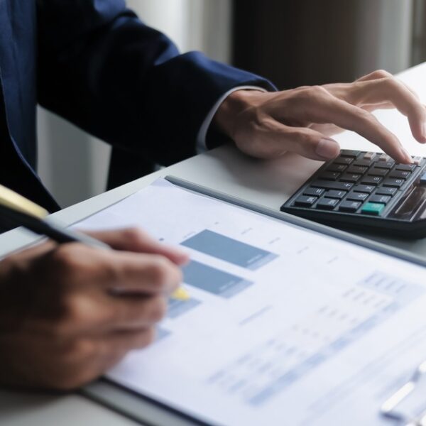 A close-up of a man in a business suit sitting at a desk, typing on a calculator, and writing on a clipboard with graphs.