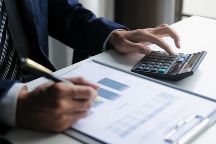A close-up of a man in a business suit sitting at a desk, typing on a calculator, and writing on a clipboard with graphs.