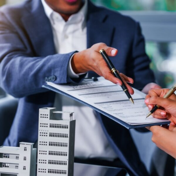 A close-up of a man handing over a clipboard of terms to a woman, both sitting in front of a multi-family real estate model.
