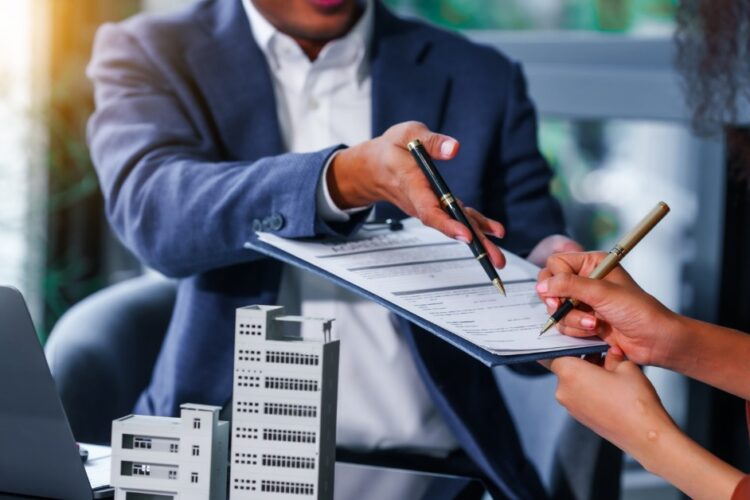 A close-up of a man handing over a clipboard of terms to a woman, both sitting in front of a multi-family real estate model.