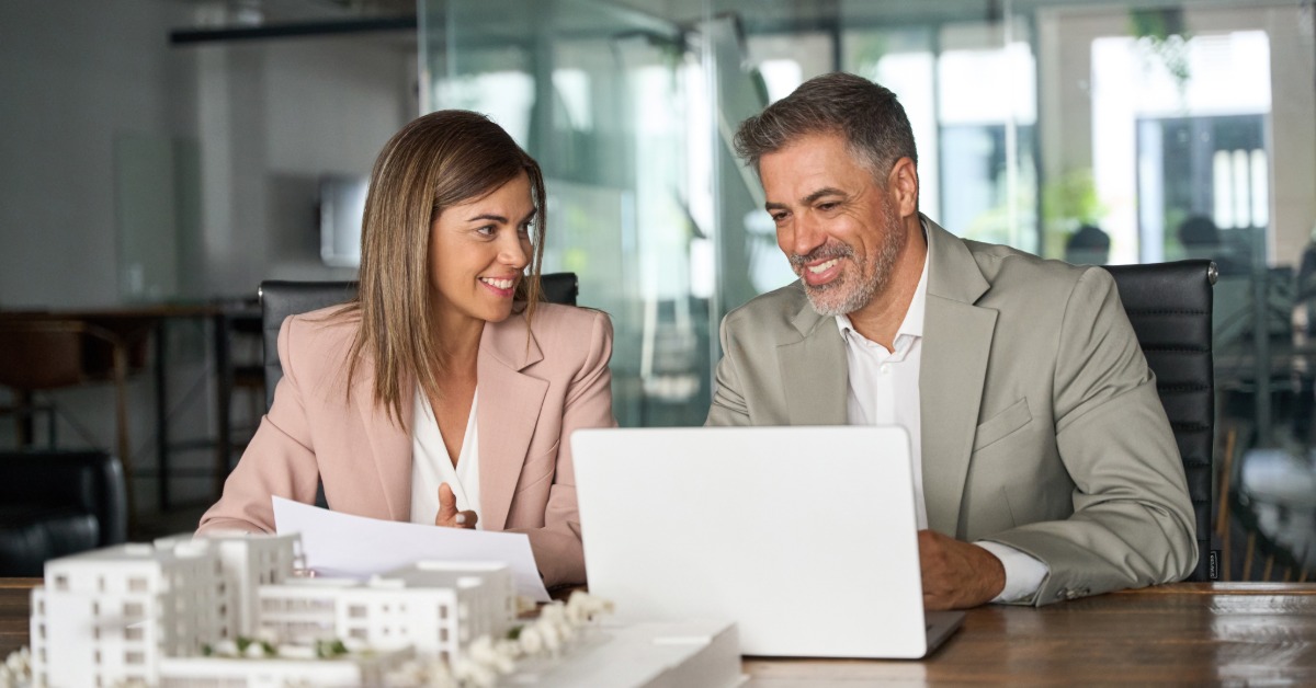 A man and a woman in business attire sitting next to each other in an office with a small-scale housing model in front of them.