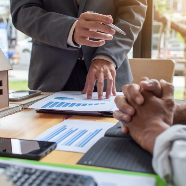 Two people in businesswear sitting at a desk with graphs and a model house sitting in front of them on the table.