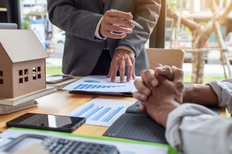 Two people in businesswear sitting at a desk with graphs and a model house sitting in front of them on the table.