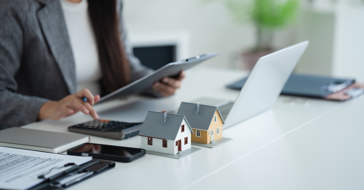 A woman dressed in business attire sits at a desk in front of two model homes with a clipboard, laptop, calculator, and documents.