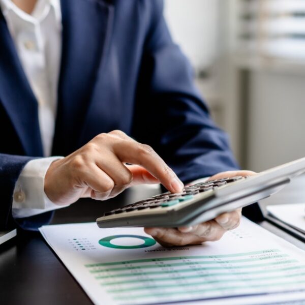 A person in a blue suit holding a calculator in one hand and typing over a clipboard and documents next to a model home.