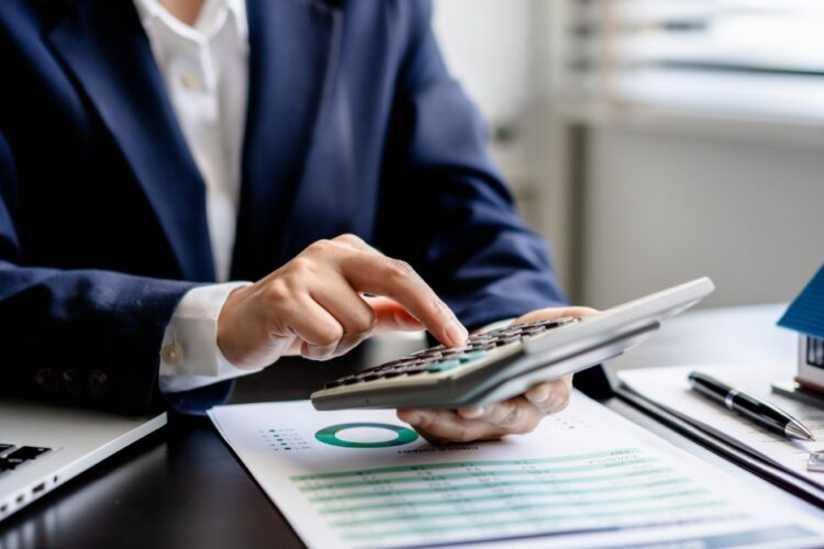 A person in a blue suit holding a calculator in one hand and typing over a clipboard and documents next to a model home.