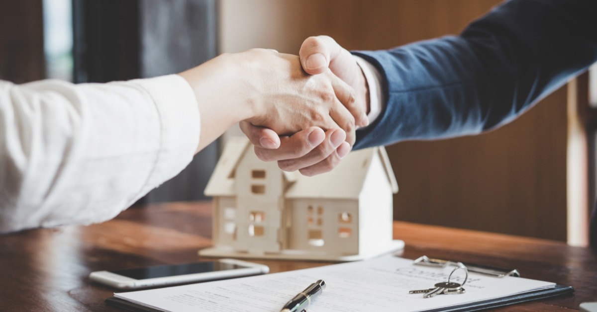 A pair of people shaking hands over a clipboard and wooden model home on an office table in an office.