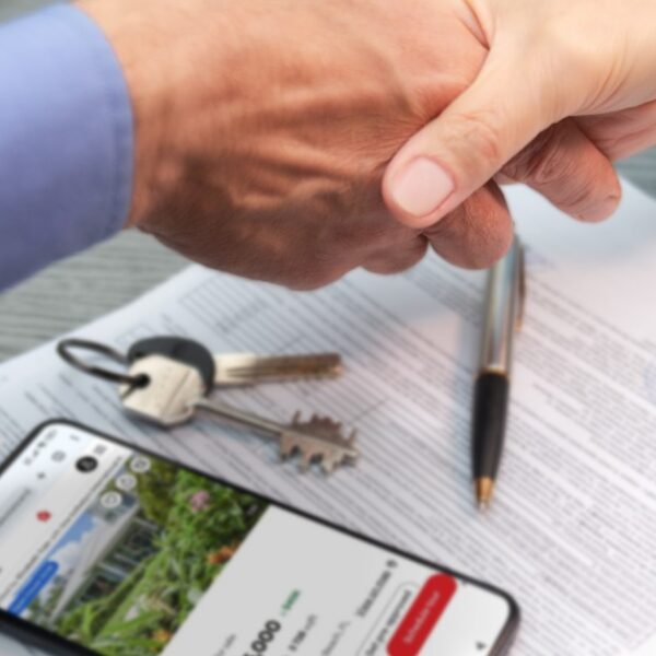 A close-up of a handshake between two people over a stack of documents, a smartphone with a home price, and a set of keys.