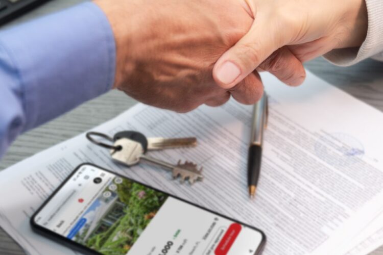 A close-up of a handshake between two people over a stack of documents, a smartphone with a home price, and a set of keys.