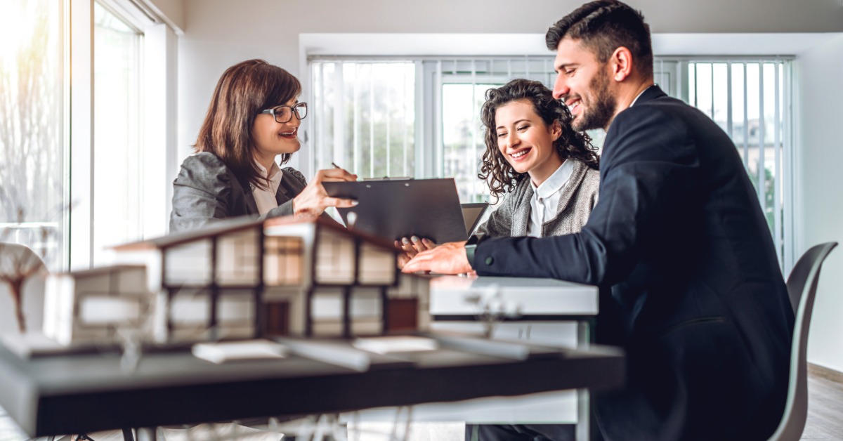 A couple sitting at a desk while a woman presents property paperwork in a bright office behind a model building.