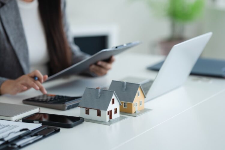 A person reviewing home loan documents beside a calculator, pen, keys, and house model on an office desk.
