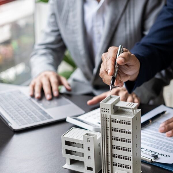 A close-up of two people in business clothing sitting at a desk with a laptop, clipboard, and a model of an office building.
