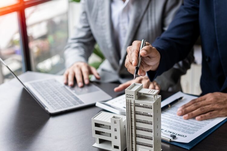 A close-up of two people in business clothing sitting at a desk with a laptop, clipboard, and a model of an office building.