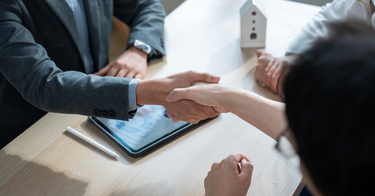 two people shaking hands across an office desk with a house model, documents, and a pen placed nearby.