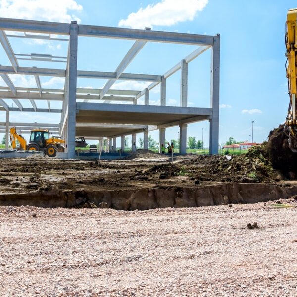 A yellow excavator digs up the land next to the framework of a building with several other heavy machines operating inside.