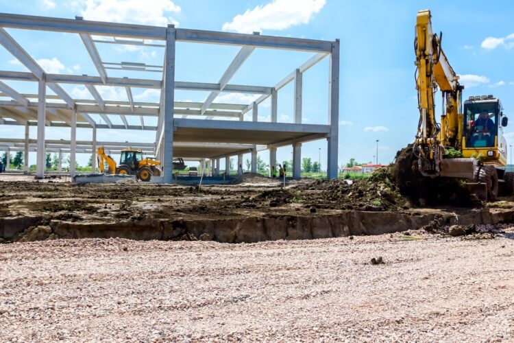 A yellow excavator digs up the land next to the framework of a building with several other heavy machines operating inside.