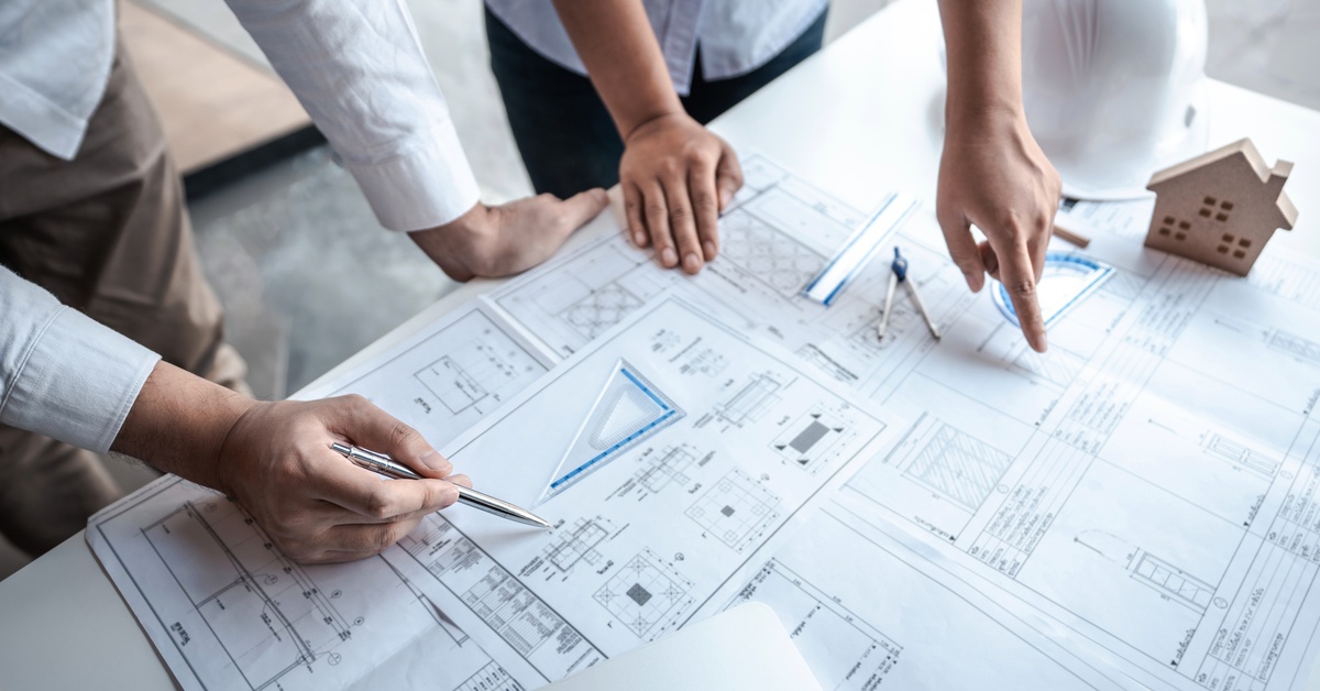 A close-up of two people leaning over a desk with various building forms and blueprints scattered across the surface.