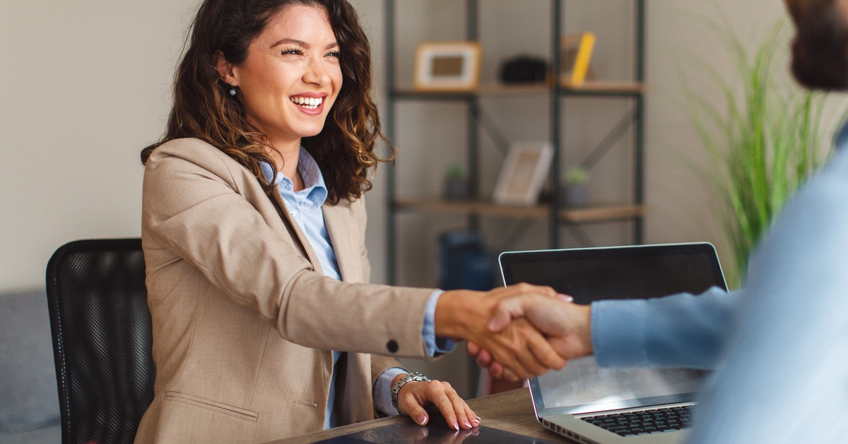 A young woman with curly dark hair wears a tan suit and shakes a man's hand across a desk with a laptop.