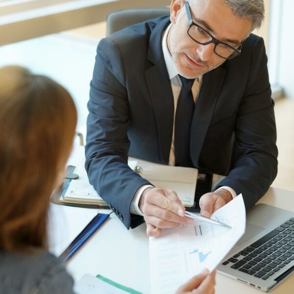 A man and a woman sitting at a desk across from each other, examining paperwork in front of a laptop.