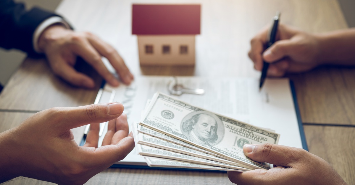 A buyer signing a deal on a clipboard while handing over a handful of one-hundred-dollar bills to a person across from them.