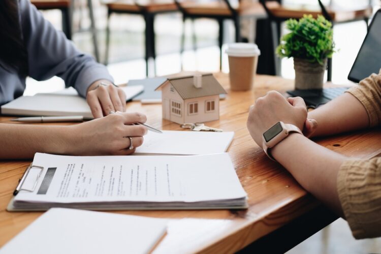 Two people sitting across from a table with a large clipboard of documents between them and a model of a home on the desk.