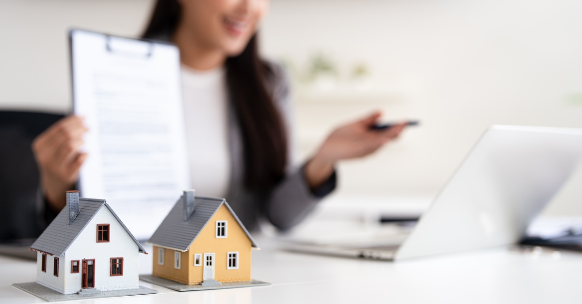 A woman holding a clipboard in front of a laptop computer, all blurred in the background behind two model houses.