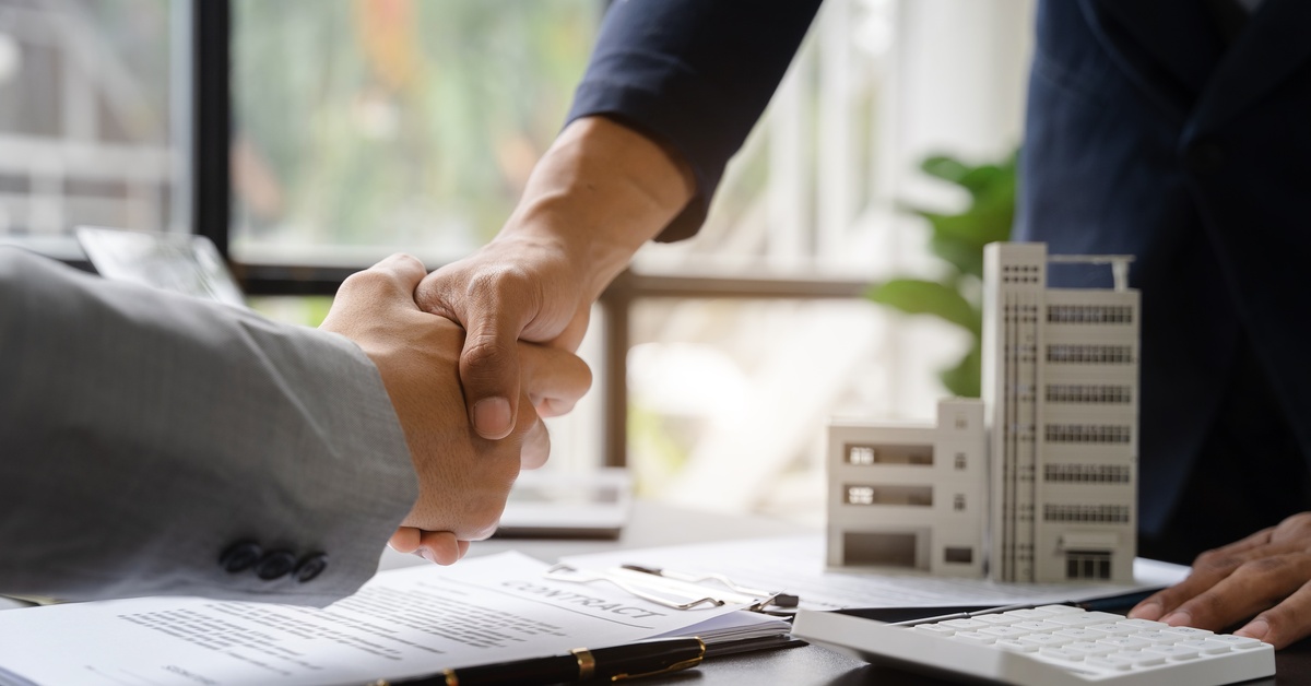 A close-up of two people shaking hands over a table with a contract and a model of a commercial building.