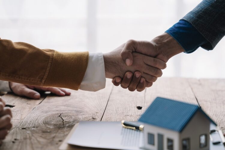 A close-up of two people shaking hands over a wooden desk with paperwork and a model house in front.