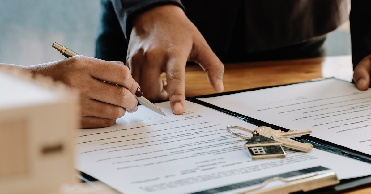 A person signing the bottom of pages of a contract with another person standing over them and a pair of house keys on the desk.