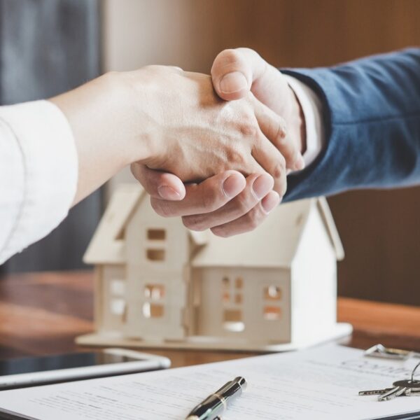 Two people wearing nice clothing reach their arms over a desk with a model home to shake hands.