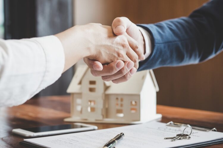 Two people wearing nice clothing reach their arms over a desk with a model home to shake hands.