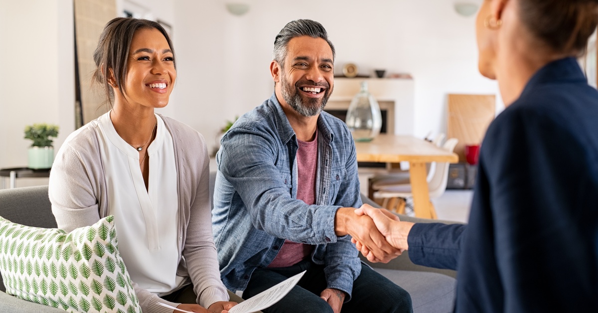 A couple sitting on a couch across from a woman in a suit, the man shaking hands with her and smiling.