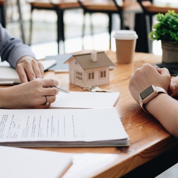 Two people sitting across from each other at a desk covered in paperwork, a model house, and an open laptop.