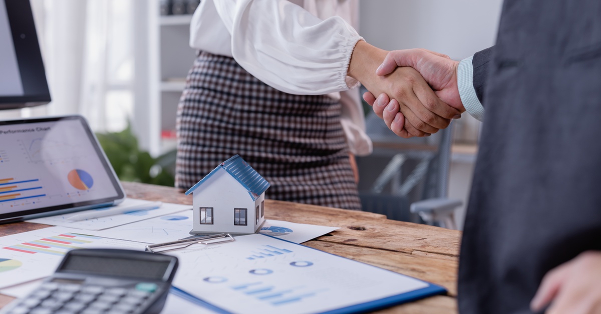 A woman in a white blouse and skirt shakes hands with a man in a suit next to a model home on a desk.