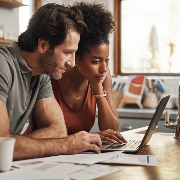 A couple lean over a kitchen countertop with various documents spread in front of them, looking at a laptop.