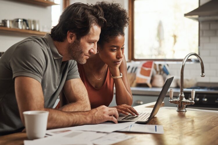 A couple lean over a kitchen countertop with various documents spread in front of them, looking at a laptop.