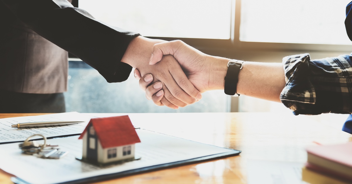 A close-up of two people shaking hands over a desk with a file, a pair of keys, and a model home sitting on it.