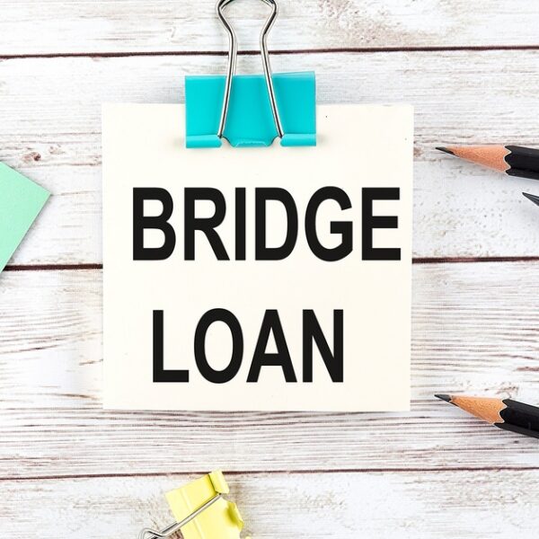 A square sheet of paper that reads "Bridge Loan" clipped with a blue clip next to black pencils and a sheet of paper.