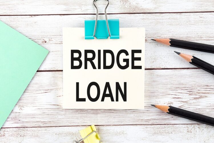 A square sheet of paper that reads "Bridge Loan" clipped with a blue clip next to black pencils and a sheet of paper.